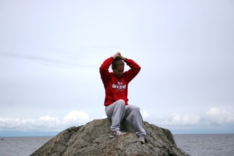 1990 Chevrolet Suburban - A person sits on a large rock overlooking the water, adjusting their hair against a cloudy sky.