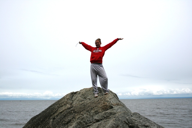 Person in red sweatshirt and grey sweatpants with arms outstretched on a large rock by the water