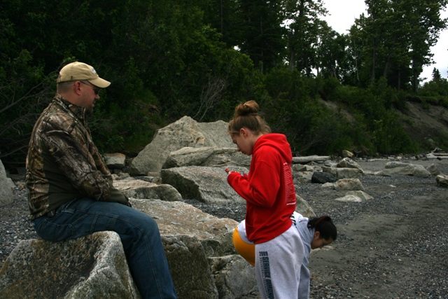 Man in camouflage jacket sits on rock, two women look at phone and ground on a rocky beach