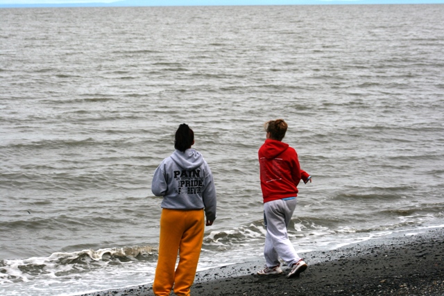 Two people stand on a dark pebble beach facing the choppy grey ocean under an overcast sky