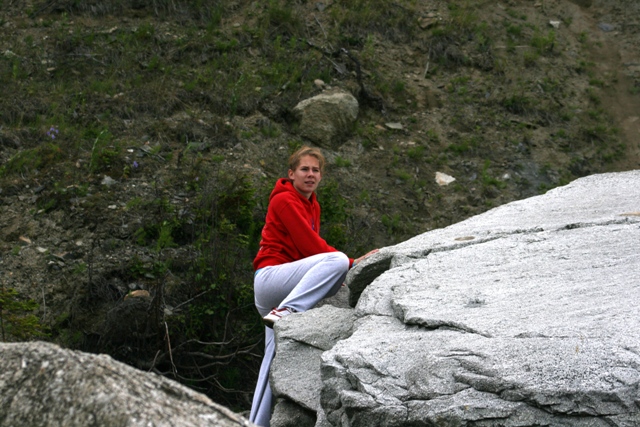 Woman in red hoodie and grey pants climbing large grey rocks with green foliage in background