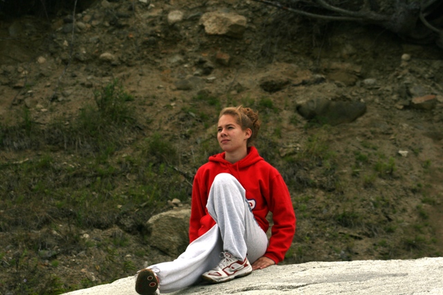 Woman in red hoodie and gray sweatpants sits on a rock, looking to the side