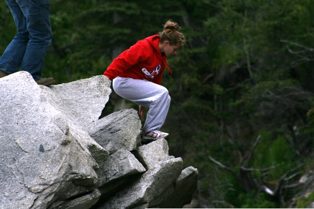 Person in red hoodie and grey sweatpants crouches on a rock ledge, another person's legs visible to the left