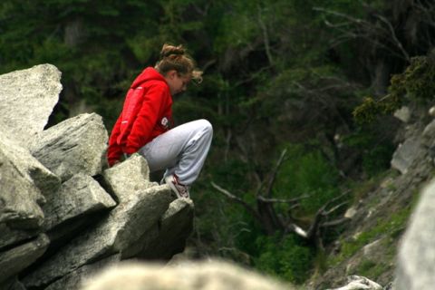 1990 Chevrolet Suburban - A person in a red hoodie and grey sweatpants sits on a rocky ledge, looking down into a wooded area.