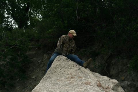1990 Chevrolet Suburban - A man wearing camouflage and a baseball cap sits on a large rock, with trees and foliage in the background.