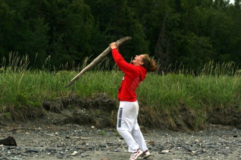 1990 Chevrolet Suburban - A young woman joyfully lifts a large piece of driftwood on a beach with tall grass and trees in the background.