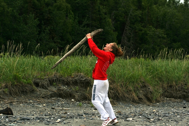 Girl in red hoodie and white pants holding a large piece of driftwood over her head on a rocky shore