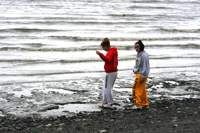 Two young women stand on a rocky shore with waves in the background