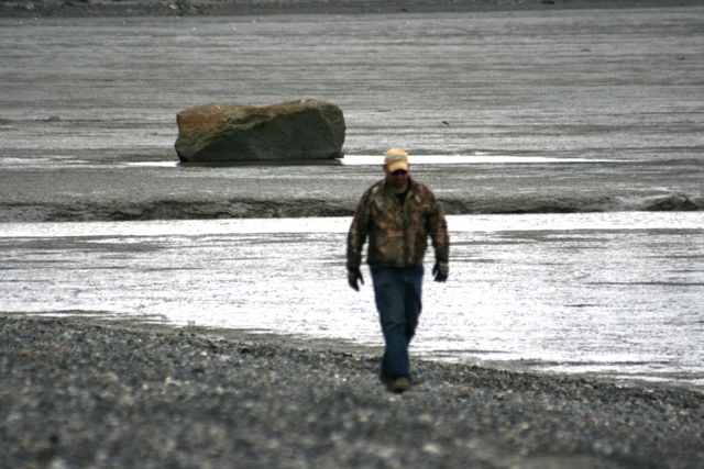 Man in camouflage jacket and jeans walks on a rocky shore with a large boulder in the background
