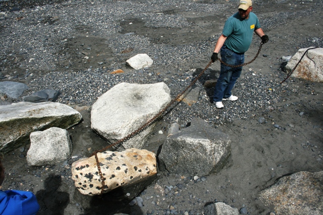 Man in a tan cap and green shirt pulls a chain attached to a large, speckled rock on a gravelly shore