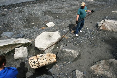 1990 Chevrolet Suburban - A man pulls a chain attached to a log on a rocky shore, with large boulders and water in the background.