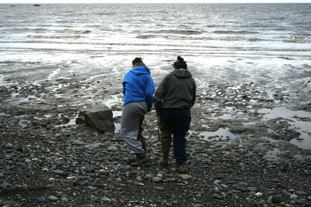 Two people stand on a rocky, muddy shore looking out at the choppy water
