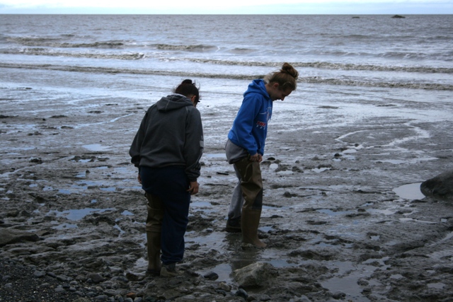 Two people wearing waders explore a muddy, rocky shore with the ocean in the background