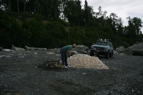1990 Chevrolet Suburban - A person works near a modified light blue SUV on a rocky shoreline with trees in the background.