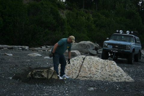 1990 Chevrolet Suburban - A man is preparing to use a winch on a light blue SUV parked on a rocky beach with trees in the background.
