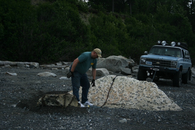 Man attaches winch cable to a rock while a light blue SUV with roof lights sits nearby on a rocky shore