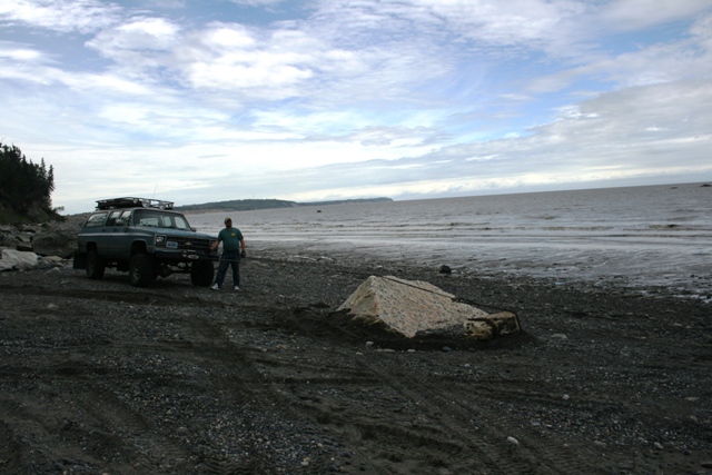 Green SUV with roof rack on a rocky beach, person standing beside it