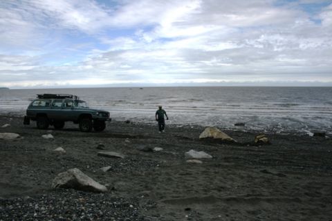 1990 Chevrolet Suburban - A teal SUV sits on a dark, rocky shore with a person walking away from it towards the water.