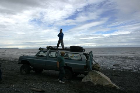 1990 Chevrolet Suburban - A person stands atop a modified SUV parked on a rocky beach with the ocean stretching to the horizon under a cloudy sky.