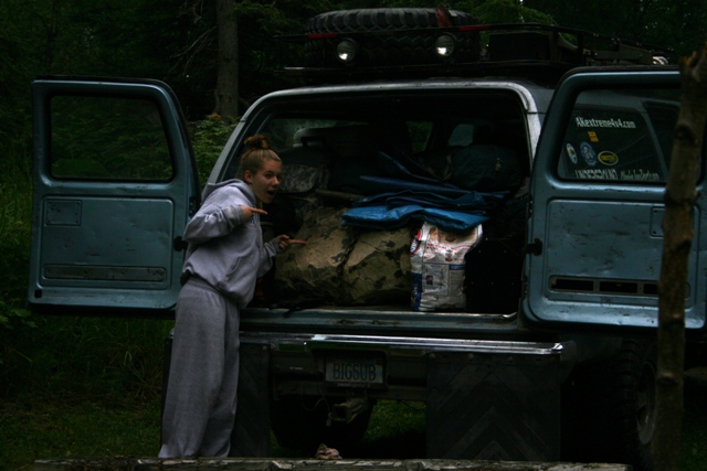 Light blue SUV with open rear doors, packed with gear. A young woman points into the cargo area. License plate reads 'BIGSUB'