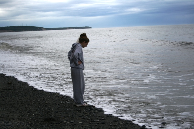 Person in a grey sweatsuit stands on a dark, pebbly beach, looking down at the incoming waves under a cloudy sky