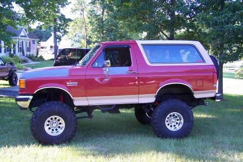 1990 Ford Bronco - A lifted red and tan Ford Bronco is parked on a grassy lawn with houses and trees in the background.