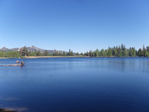 1991 Ford Bronco - A calm lake reflects the clear blue sky, with a treeline and snow-capped mountains in the distance.
