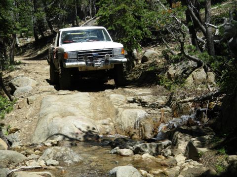 1991 Ford Bronco - White Ford F-Series truck navigates a challenging rocky terrain on a wooded trail next to a flowing stream.