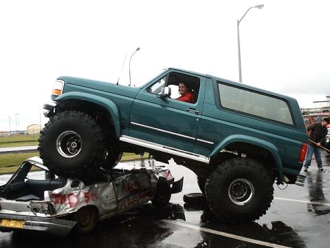 1996 Ford Bronco - A green 1996 Ford Bronco is shown parked on top of a heavily damaged car, with a driver visible inside the Bronco.