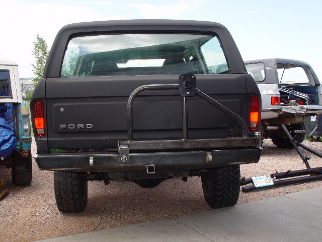 Rear view of a black Ford Bronco with a custom bumper and spare tire mount. Another vehicle is parked behind it