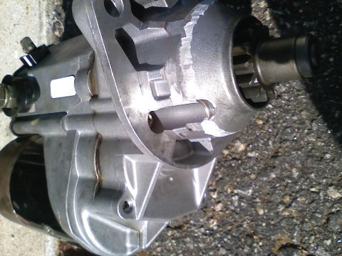 1990 Ford Bronco - Close-up of a removed starter motor lying on asphalt, showing the cast-aluminum gear-reduction nose housing, the splined pinion drive gear…