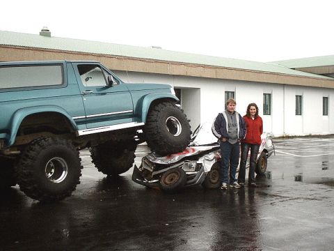 1996 Ford Bronco - A large lifted truck is parked on top of a severely damaged car, with two people standing to the side.