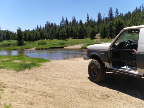 1991 Ford Bronco - 1991 Ford Bronco parked on a sandy bank next to a river with a wooded area behind it.