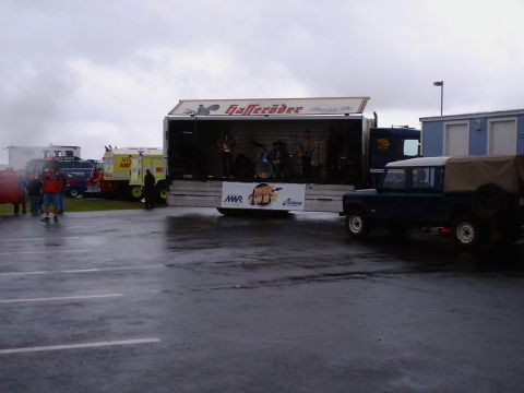 1996 Ford Bronco - A band performs on a stage set up on the back of a truck, with a Land Rover parked nearby.