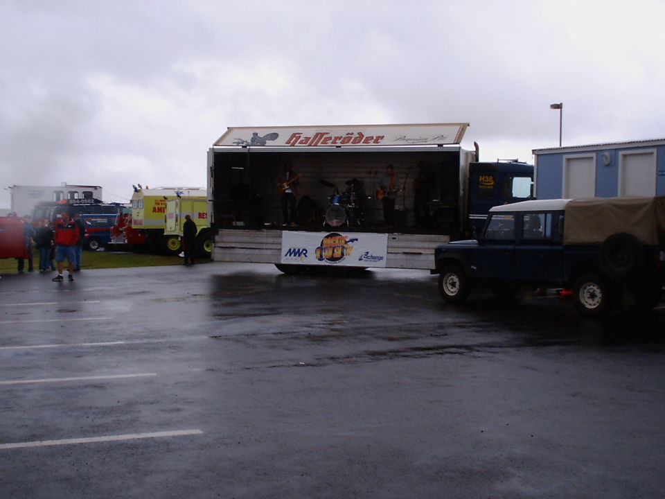 Wet outdoor lot with a band on a stage truck, emergency vehicles, and a blue Land Rover Defender
