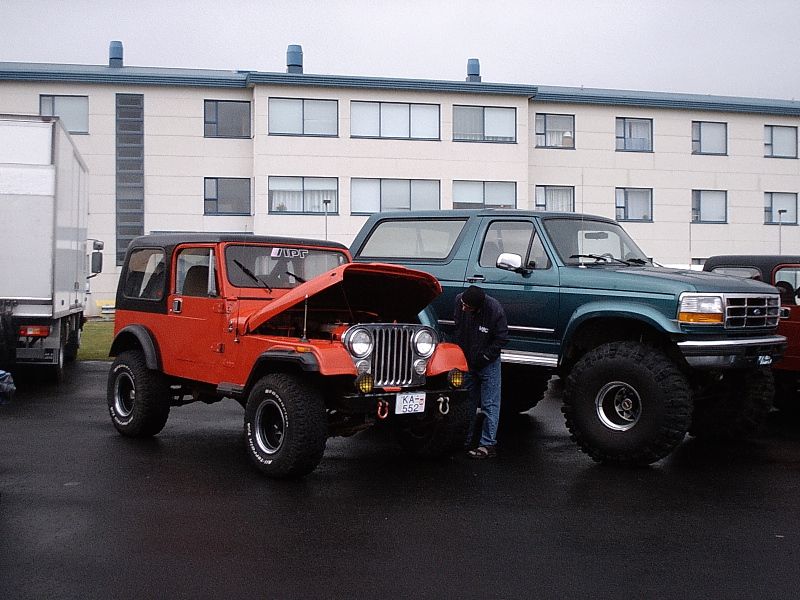 Orange Jeep with hood up next to green lifted Ford Bronco in wet lot