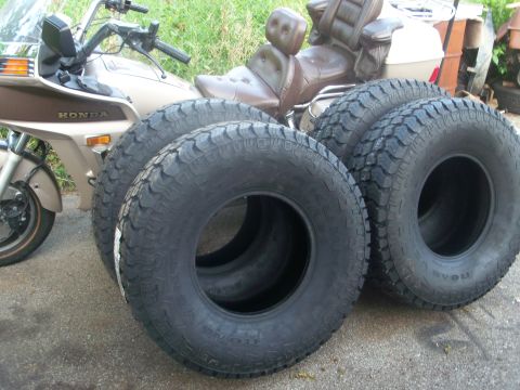 1989 Ford Bronco - Four large off-road tires are displayed in front of a motorcycle, with a portion of a building visible in the background.