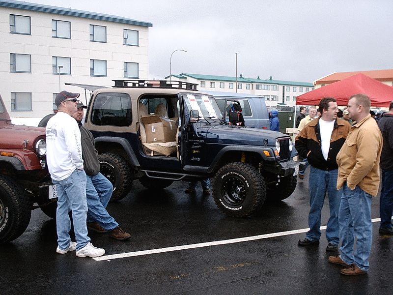 Lifted dark blue Jeep Wrangler on mud tires at a wet outdoor car meet with people around it