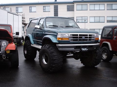 1996 Ford Bronco - A modified Ford Bronco sits on a wet asphalt lot next to other vehicles.