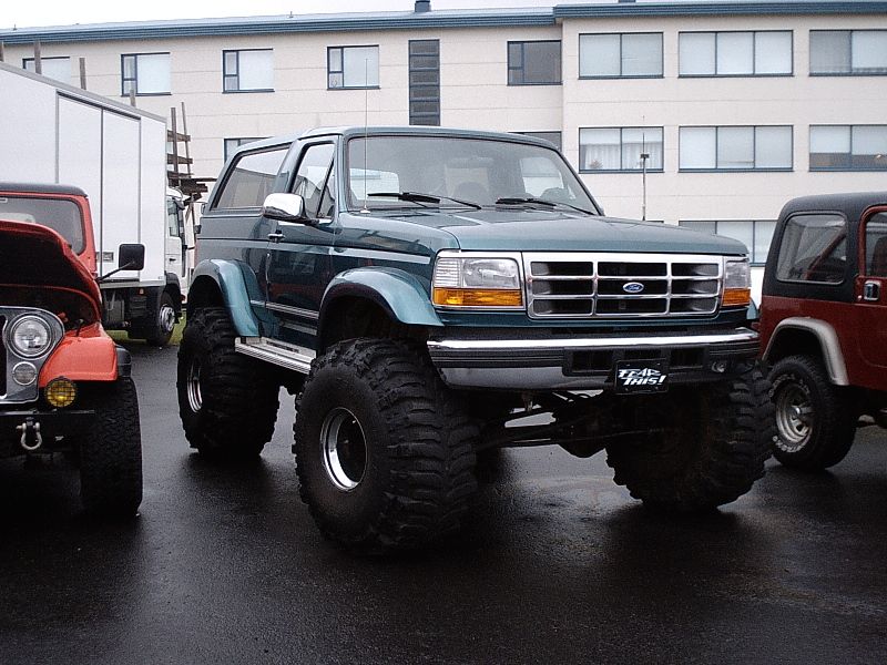 Lifted teal Ford Bronco on big mud tires in wet lot, flanked by red Jeeps.