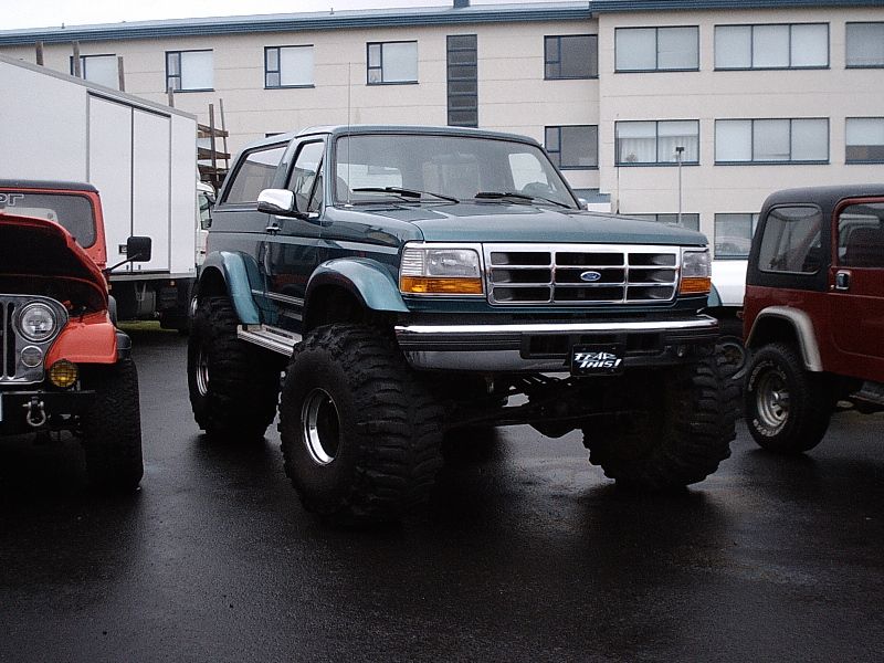 Lifted teal Ford Bronco on huge mud tires, front 3/4 view, between two red Jeeps