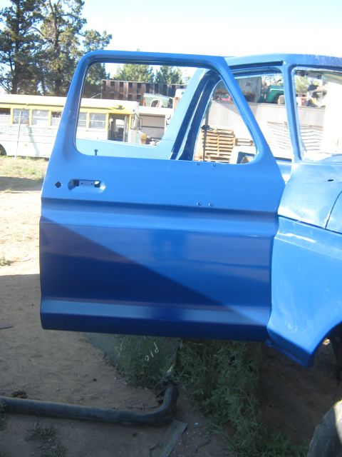 1978 Ford Bronco - A blue pickup truck door is open, revealing the interior frame. Other vehicles are visible in the background.