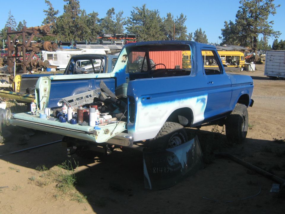 Blue 1978 Ford Bronco in a junkyard, undergoing repairs, with its tailgate down and parts scattered