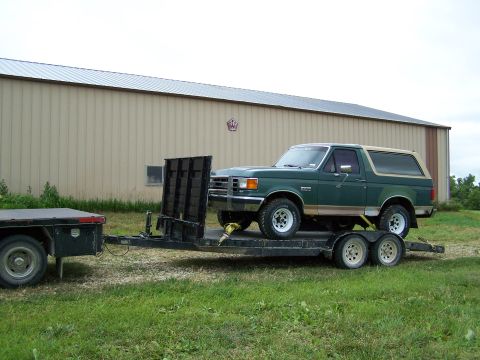 1989 Ford Bronco - A green Ford Bronco is being transported on a trailer, parked in front of a large industrial building.