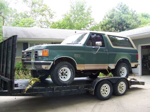 1989 Ford Bronco - A green 1987 Ford Bronco is loaded onto a trailer, showing its front-left side with tan trim and white wheels.