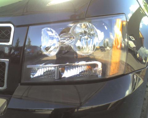 1993 Ford C-350 (Centurion Conversions) - Front headlight assembly of a dark vehicle, reflecting the sky and surrounding environment.