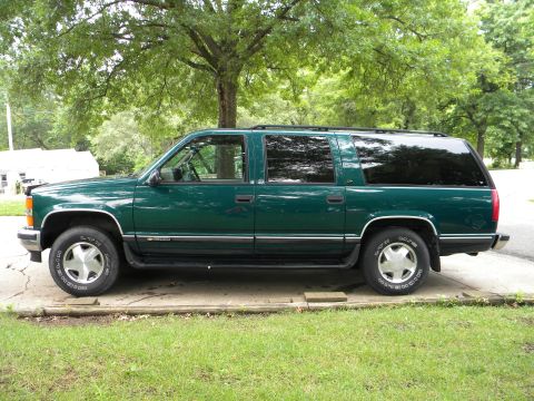 1999 Chevrolet Suburban - This green SUV is parked on a concrete pad with grass and trees in the background.