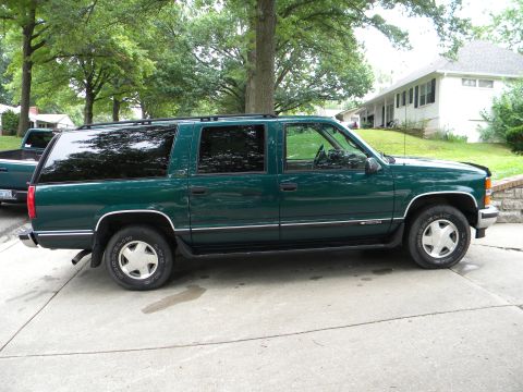 1999 Chevrolet Suburban - A green Chevrolet Suburban is parked on a concrete driveway next to a pickup truck.