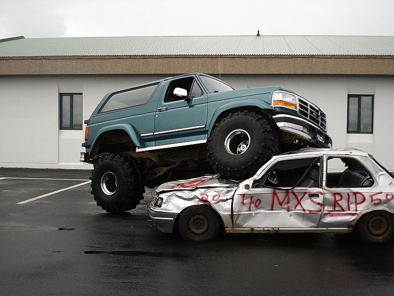 Teal lifted Ford Bronco parked on top of a crushed silver sedan with red graffiti.