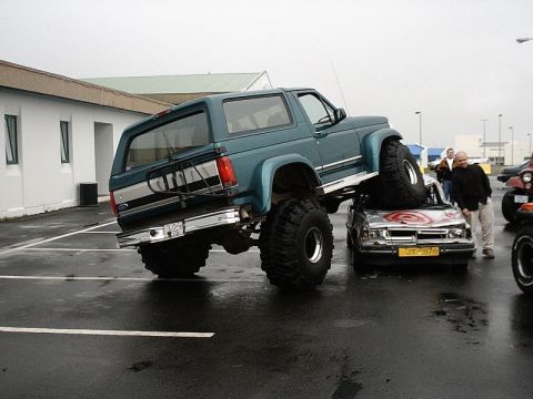 1996 Ford Bronco - A heavily modified Ford Bronco is parked on top of a sedan, crushing its roof.
